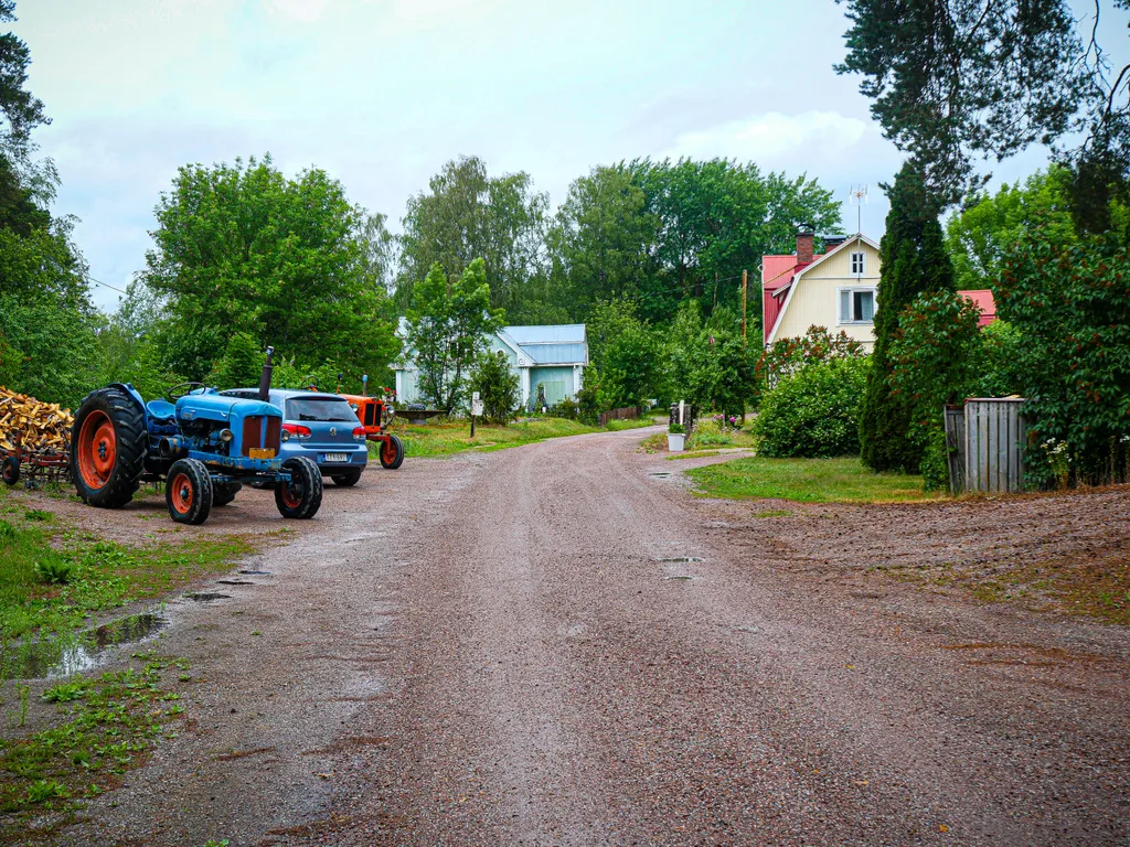 Eine Schotterstraße führt durch einen Bauernhof. Neben der Straße parken ein alter Traktor und ein Auto.