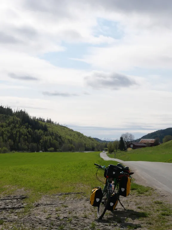 Mein Fahrrad steht am Straßenrand auf einer grünen Wieße. Im Hintergrund ein Bauernhof und Wald. Der Himmel ist bewölkt.