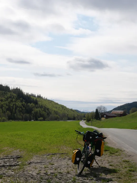 Mein Fahrrad steht am Straßenrand auf einer grünen Wieße. Im Hintergrund ein Bauernhof und Wald. Der Himmel ist bewölkt.