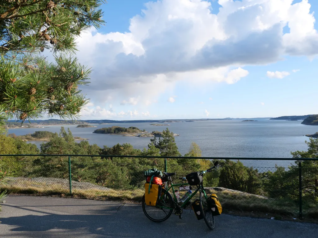 Mein Fahrrad steht auf einer Anhöhe auf einem Radweg vor einem Zaun. Dahinter sieht man einen Ausblick auf kleinere Felsinseln und eines Fjords.