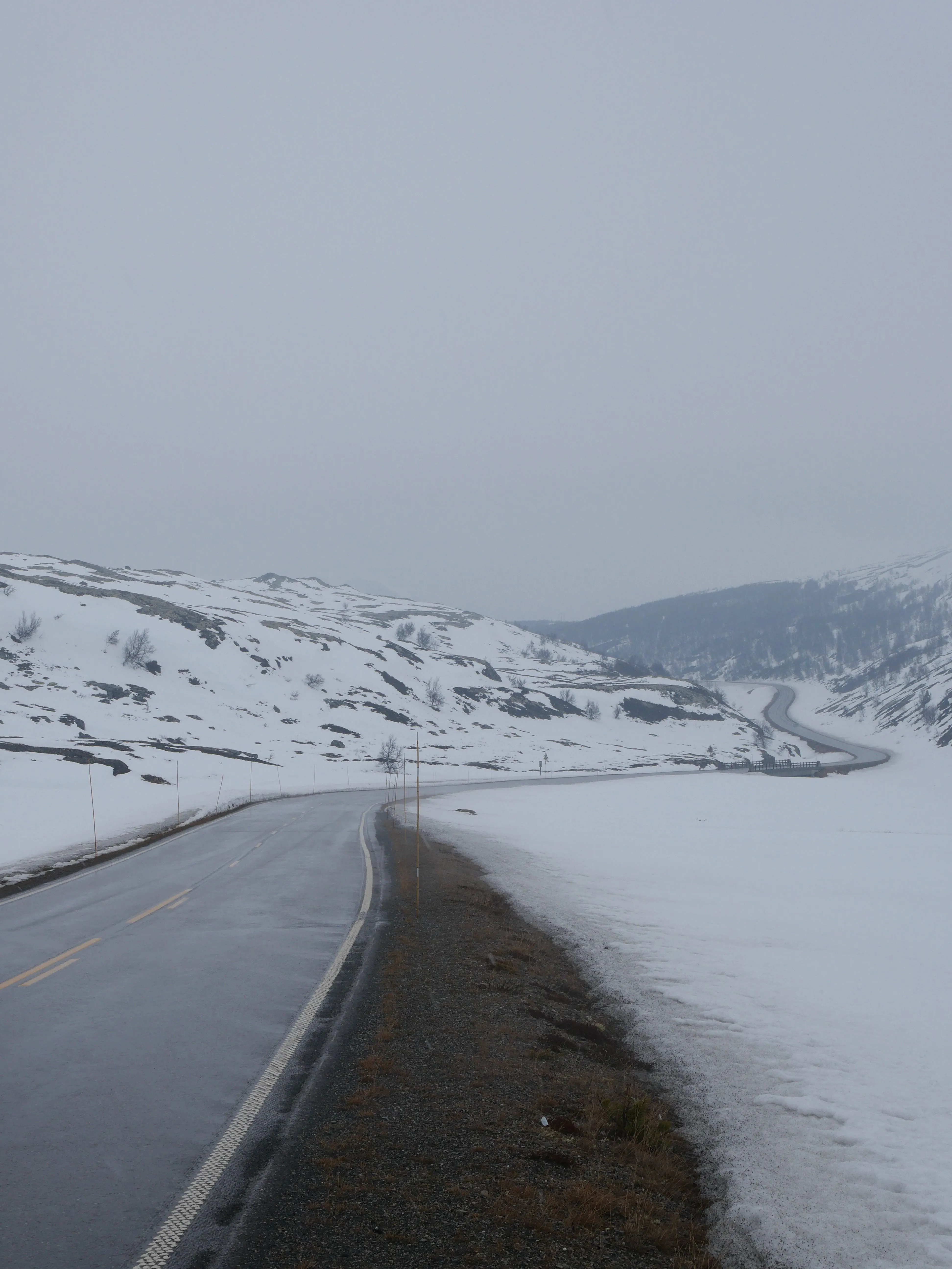 Eine vom Regen nasse kurvenreiche Straße zwischen Schneebedeckten Hügeln. Die Wolken sind niedrig und der Himmel ist grau.