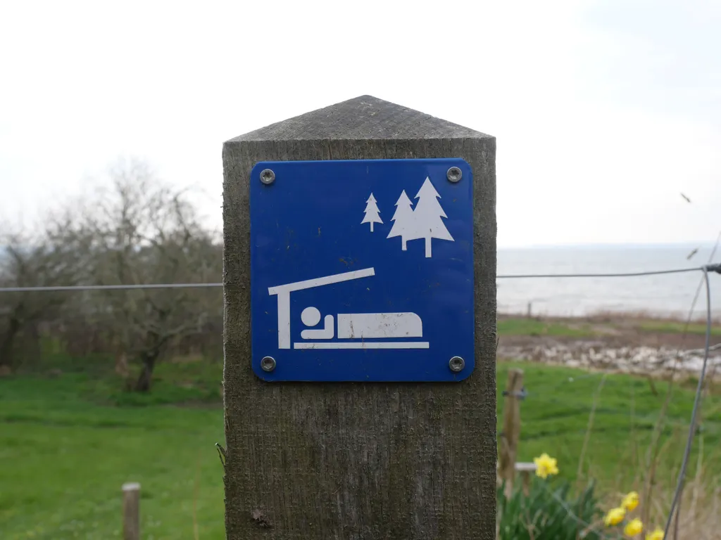 Ein Schild das auf einen Shelter hinweist. Im Hintergrund die dänische Küste mit einer grünen Wiese.