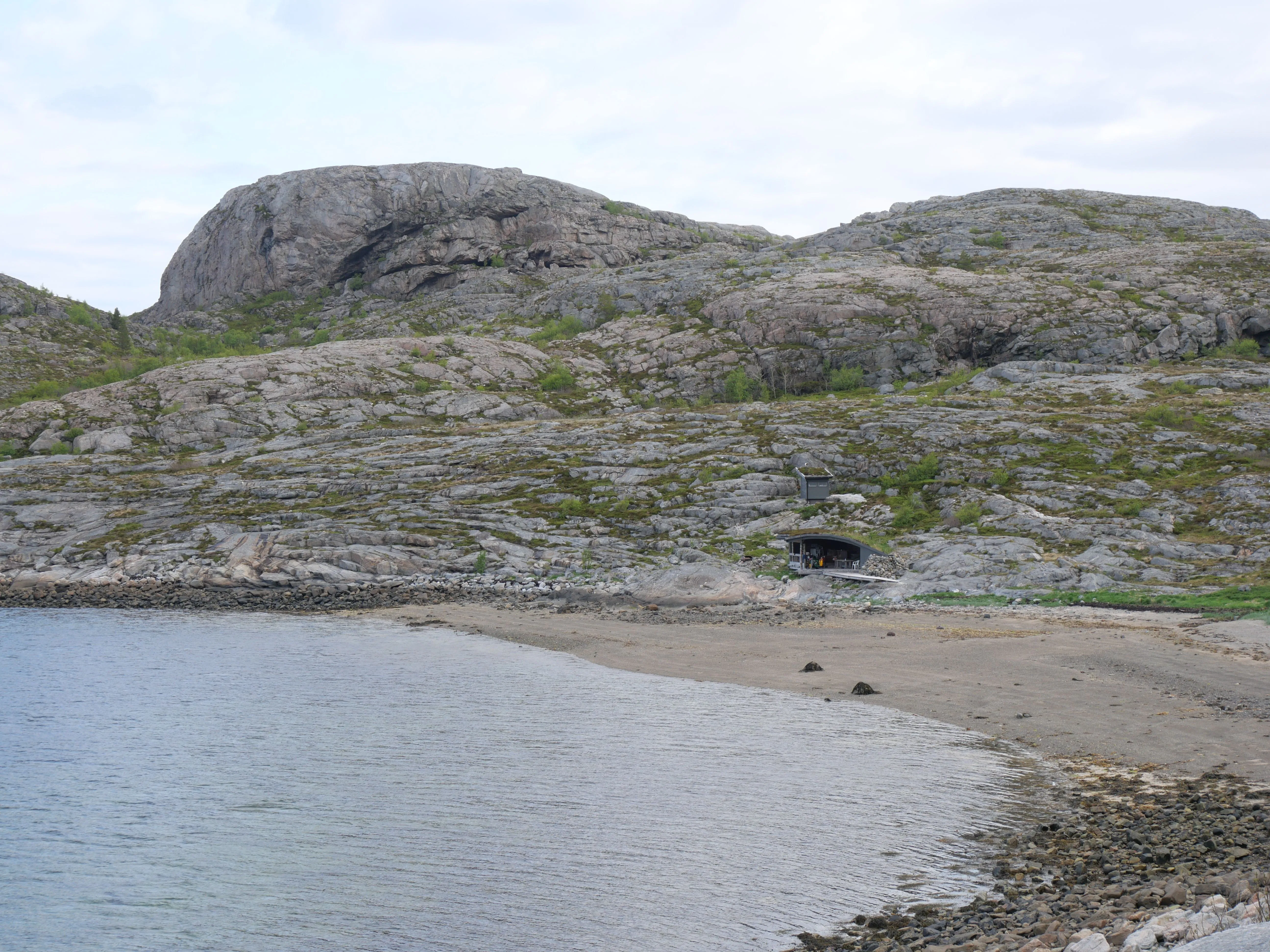 Steinige Hügellandschaft direkt am Sandsstrand. Dazwischen ganz klein noch zu sehen eine kleine offene Schutzhütte.