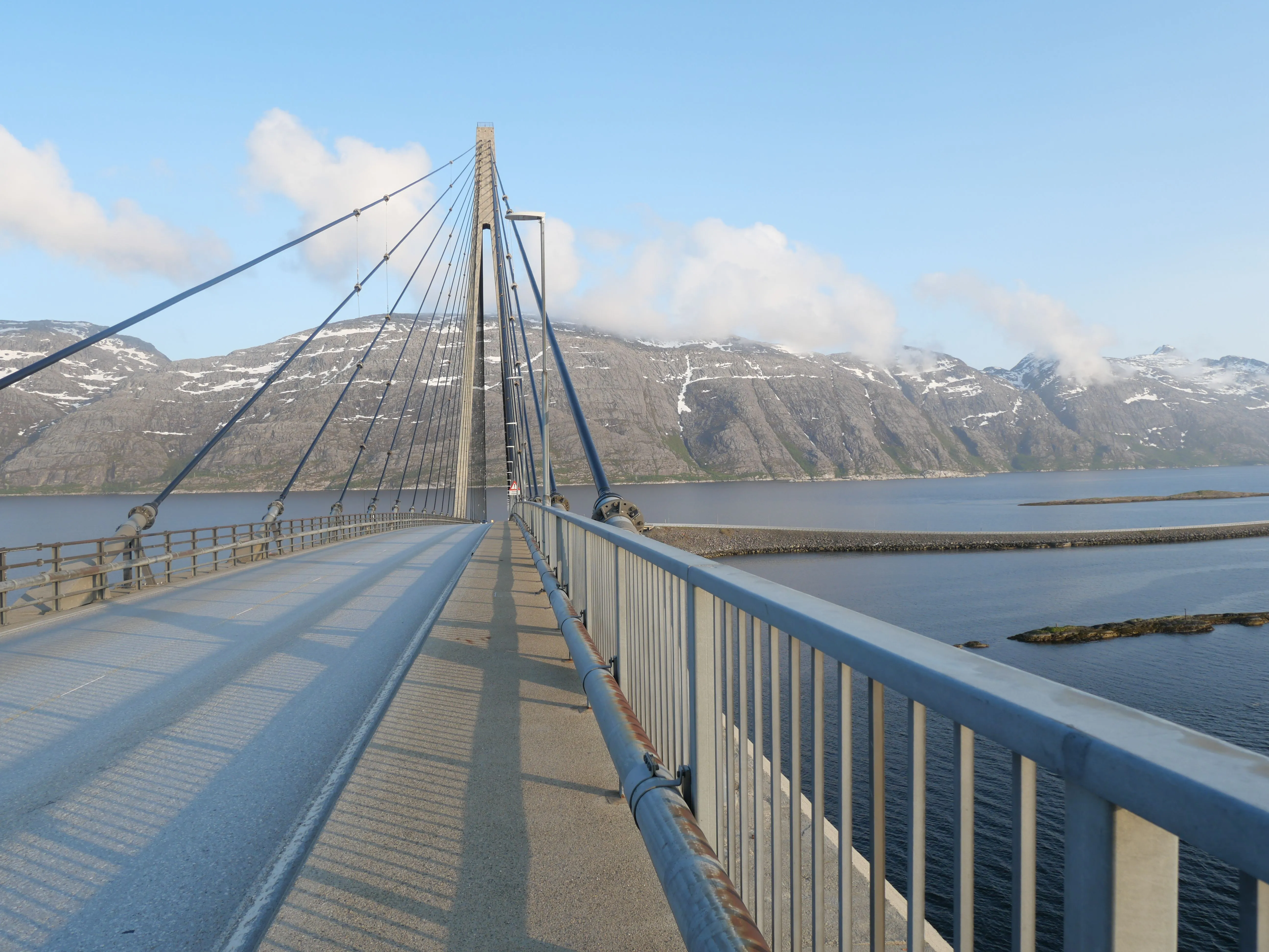 Blick gerade auf eine große Stahlbrücke über Wasser. Im Hintergrund sind Gebirge zu sehen