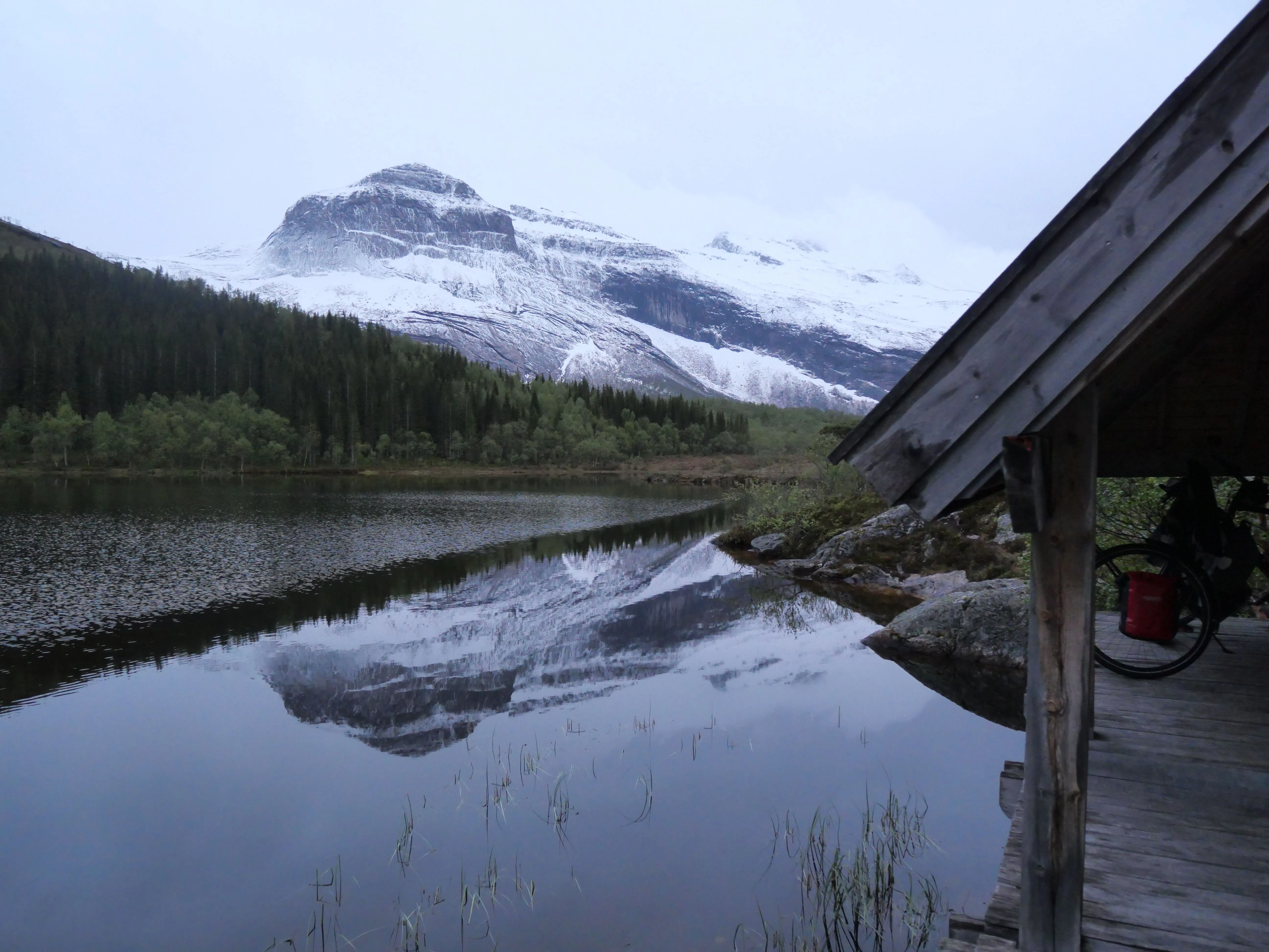 Ein Schneebedeckter Berg spiegelt sich in einem See. Das Foto wurde von einem überdachten Steg aufgenommen, welcher am Rand noch zu sehen ist.