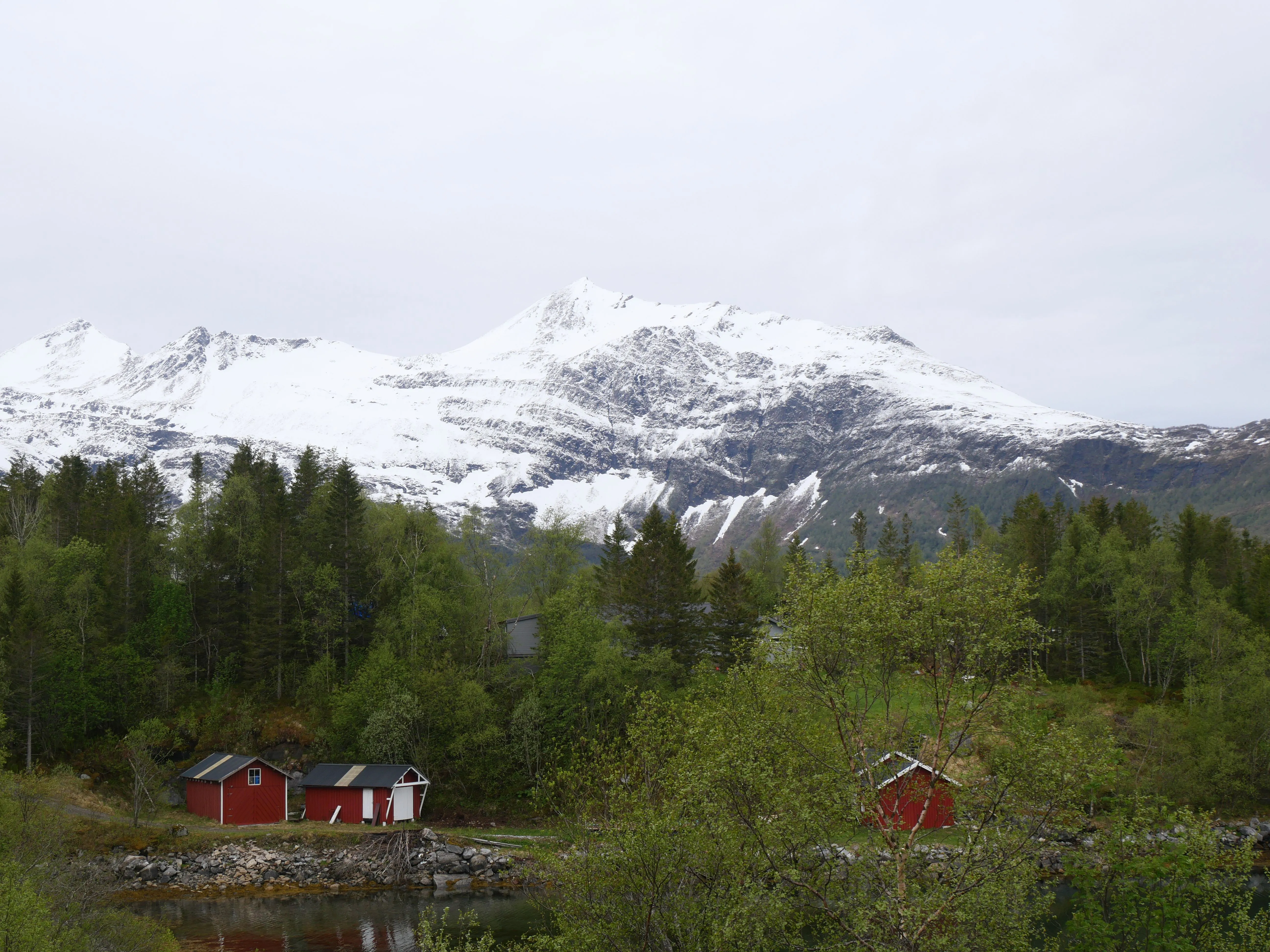 Rote Häuser sind am Waldesrand zu sehen. Dahinter ragen schneebedckte Berggipfel in den Himmel