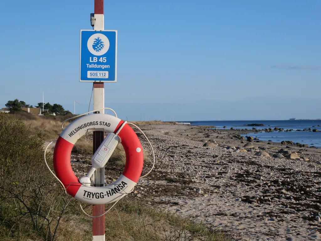 Ein rot-weißer Rettungsring ist an einem rot-weißen Holzpfahl angebracht. Der Pfahl steht am Strand. Man sieht ein wenig Gras, Sand mit Steinen und dann das Meerwasser.