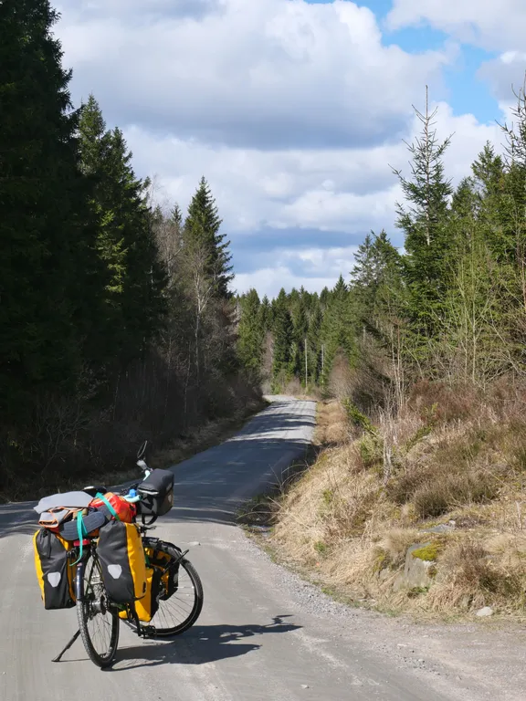 Mein Fahrrad steht auf einer ungeteerten Straße zwischen Nadelbäumen. Am Himmel sind einige Wolken zu sehne durch welche teilweise blauer Himmel durchblitzt.